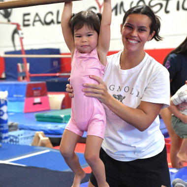 Child in a pink leotard hangs from a gymnastics bar as a smiling instructor supports her in a gymnasium mission.